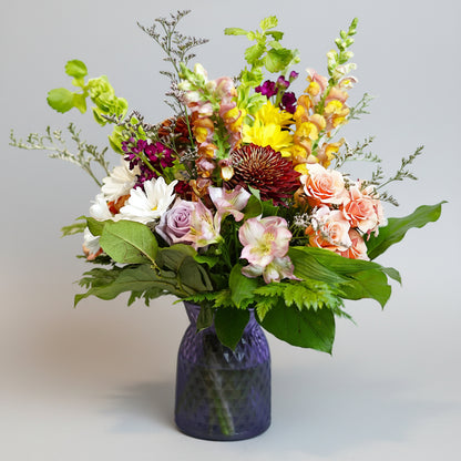 A vibrant mixed floral arrangement in a textured purple glass vase, featuring pink, yellow, white, and purple flowers with lush green foliage, set against a plain light background.
