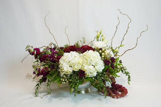A floral arrangement in a clear glass vase featuring white hydrangeas, deep burgundy roses, purple stock flowers, green foliage, and delicate bare branches, set against a plain light background.